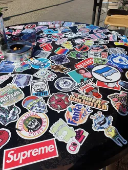 Street sticker This image shows a table covered in a variety of stickers.  The stickers include various popular brands, cartoon characters, and designs.  A Supreme sticker is prominently featured in the bottom center.
