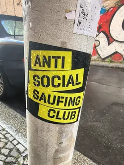Street sticker A black sticker featuring wavy yellow blocks with the stencil-style text 'ANTI SOCIAL SAUFING CLUB'. It is wet with rain droplets and stuck to a textured pole.