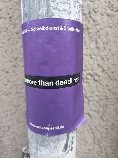 This sticker is primarily purple with a black banner displaying the message "Life is more than deadlines" in white text. The website address is printed at the bottom. The sticker is placed on a pole.
