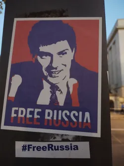 Street sticker A sticker featuring a stylized portrait of a man, reminiscent of Soviet-era propaganda posters, with the text "FREE RUSSIA" prominently displayed. The colors are primarily red, white, and blue, evoking the Russian flag. A smaller sticker below reads "#FreeRussia".