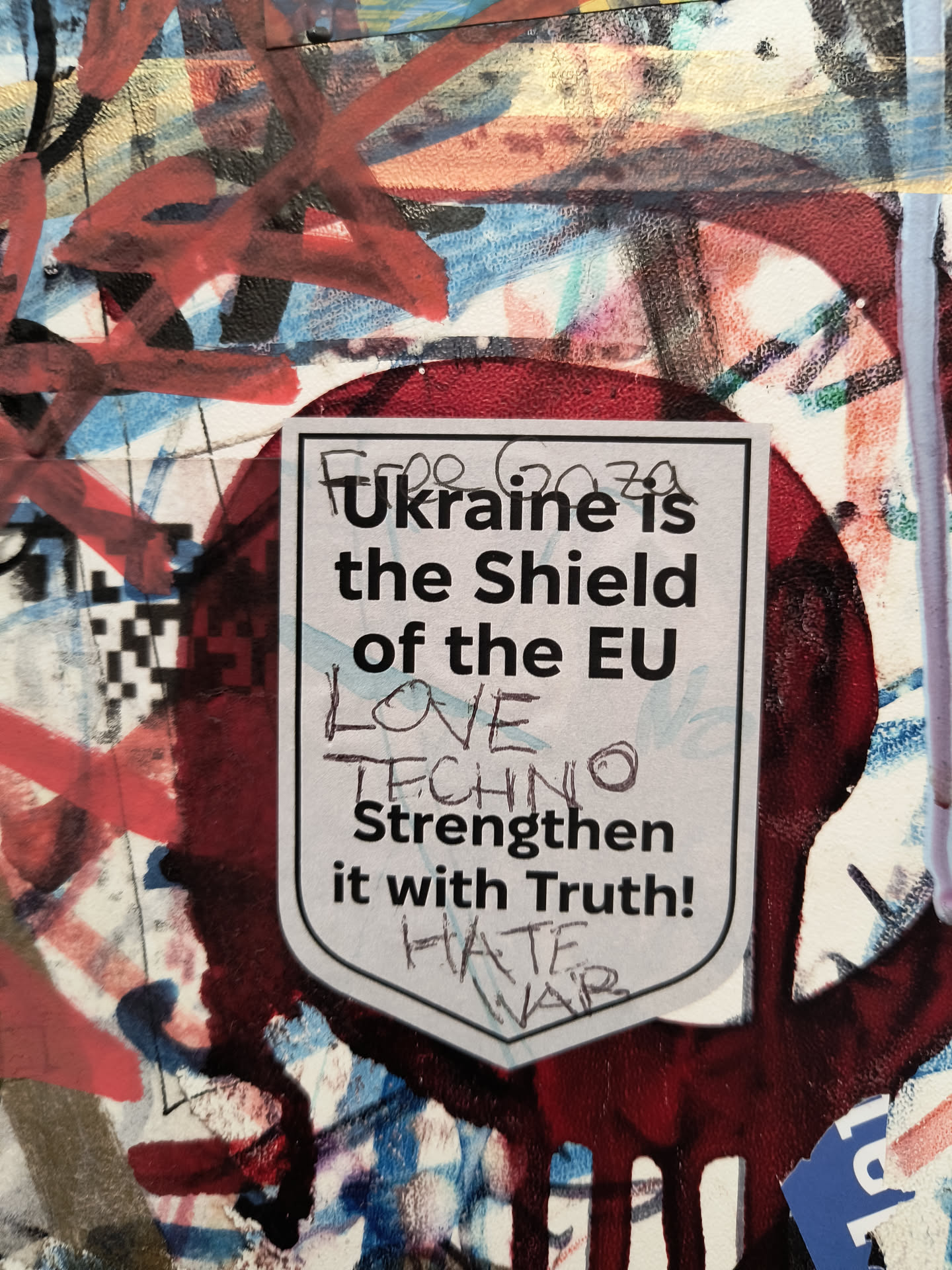 The sticker is shield-shaped and features text on a light grey background with a dark border. The text reads 'Ukraine is the Shield of the EU LOVE TECHNO Strengthen it with Truth! HATE WAR'. The top of the shield has what appears to be a handwritten 'Free Gaza 2020'. The sticker is placed on a surface covered in various forms of graffiti and other pasted materials.