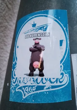 A blue and white sticker featuring a cartoon cow with its arms crossed.  The text on the sticker says "DU HURENSOHN" which is a very vulgar German insult. There is also a ship graphic and the year 1965. The style is reminiscent of vintage or retro designs.