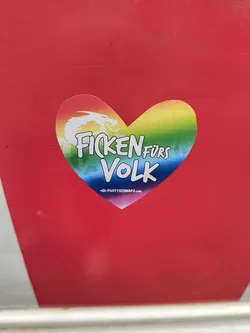 Street sticker Heart-shaped sticker with a rainbow design, a dragon, and the text "Ficken fürs Volk."  The text is written in German and translates roughly to "Fuck for the People". A website address is also visible at the bottom.