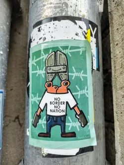 Street sticker A sticker featuring an illustration of a character with a pair of wire cutters for a head, wearing a shirt that says "NO BORDER NO NATION." The character is standing in front of a barbed wire fence. The style is cartoonish and slightly edgy.