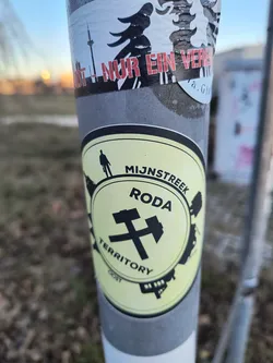 The sticker is an oval-shaped design with a light yellow-green background and black elements. It features a central image of two crossed mining hammers, a classic symbol of coal mining. Arcing above the hammers is the word 'MIJNSTREEK', and below them, 'TERRITORY'. The word 'RODA' is prominently displayed in the center. Along the top inner edge of the oval, a small black silhouette of a person walking is visible. The outer edge of the sticker is bordered by black silhouettes of industrial structures and buildings, suggesting a cityscape or mining infrastructure. A small 'OOST' is visible at the very bottom.