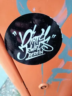 Street sticker Round black and white sticker with the text "Dirty Jewels" in a stylized graffiti font and "Berlin" written in a smaller, simpler font underneath. The sticker is applied to an orange surface with some other graffiti markings visible around it.