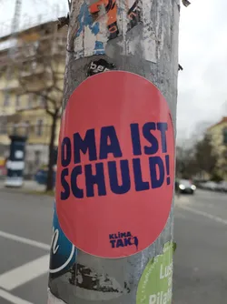 Street sticker A round pink sticker with the text "OMA IST SCHULD!" in dark blue letters.  At the bottom, a small logo says "Klima Tak". The sticker is affixed to a pole covered in other stickers.