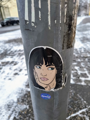 A sticker featuring a stylized illustration of a woman with dark hair, resting her chin on her hand. The art style is reminiscent of pop art or comic book aesthetics. There's a small, round logo underneath that reads ' xenia ' in stylized lettering. The sticker is affixed to a weathered metal pole, possibly in an urban environment. There's snow on the ground, suggesting a winter setting.