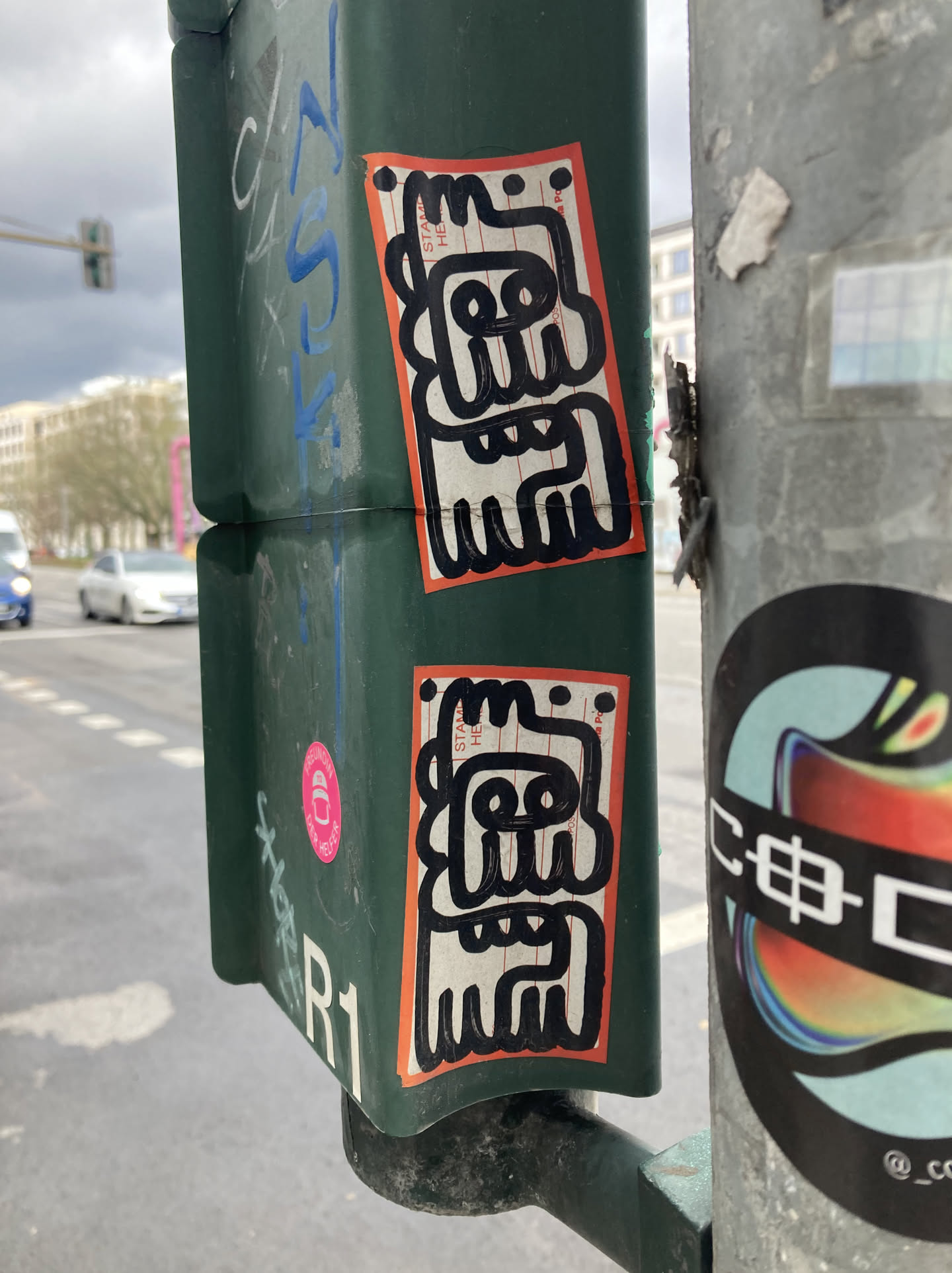 The central focus of the image features two identical stickers, each with an orange border. Inside the border, the sticker has a white background with vertical lines. On this background, a black abstract character is drawn with thick, bold lines. The character has a square-like head with circular eyes and a series of jagged lines forming a mouth and body. The overall style is reminiscent of a cartoon or doodle.