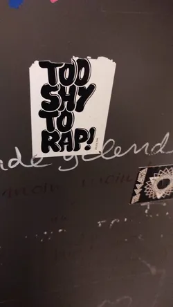 A sticker with the text "Too shy to rap!" in a stylized font. The text is black on a white background. The sticker is slightly worn around the edges. 