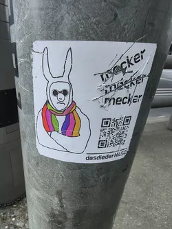 Street sticker A white sticker featuring a stylized, somewhat minimalist drawing of a figure wearing a white bunny costume or mask with long ears. The face inside the mask has a simple, grumpy expression with a downturned mouth and round, dark sunglasses. The figure's arms are crossed over its chest. A prominent feature is a brightly colored rainbow-striped scarf wrapped around its neck, displaying vibrant bands of red, orange, yellow, green, blue, and purple. To the right of the bunny, there is black text that appears to be scratched out or crossed through, reading 'meckern' multiple times. Below this text is a QR code, and at the very bottom of the sticker, the text 'dasdiederHASE' is visible. The sticker itself shows signs of wear, including creases and slight peeling, indicating it's a physical sticker applied to a textured grey pole.