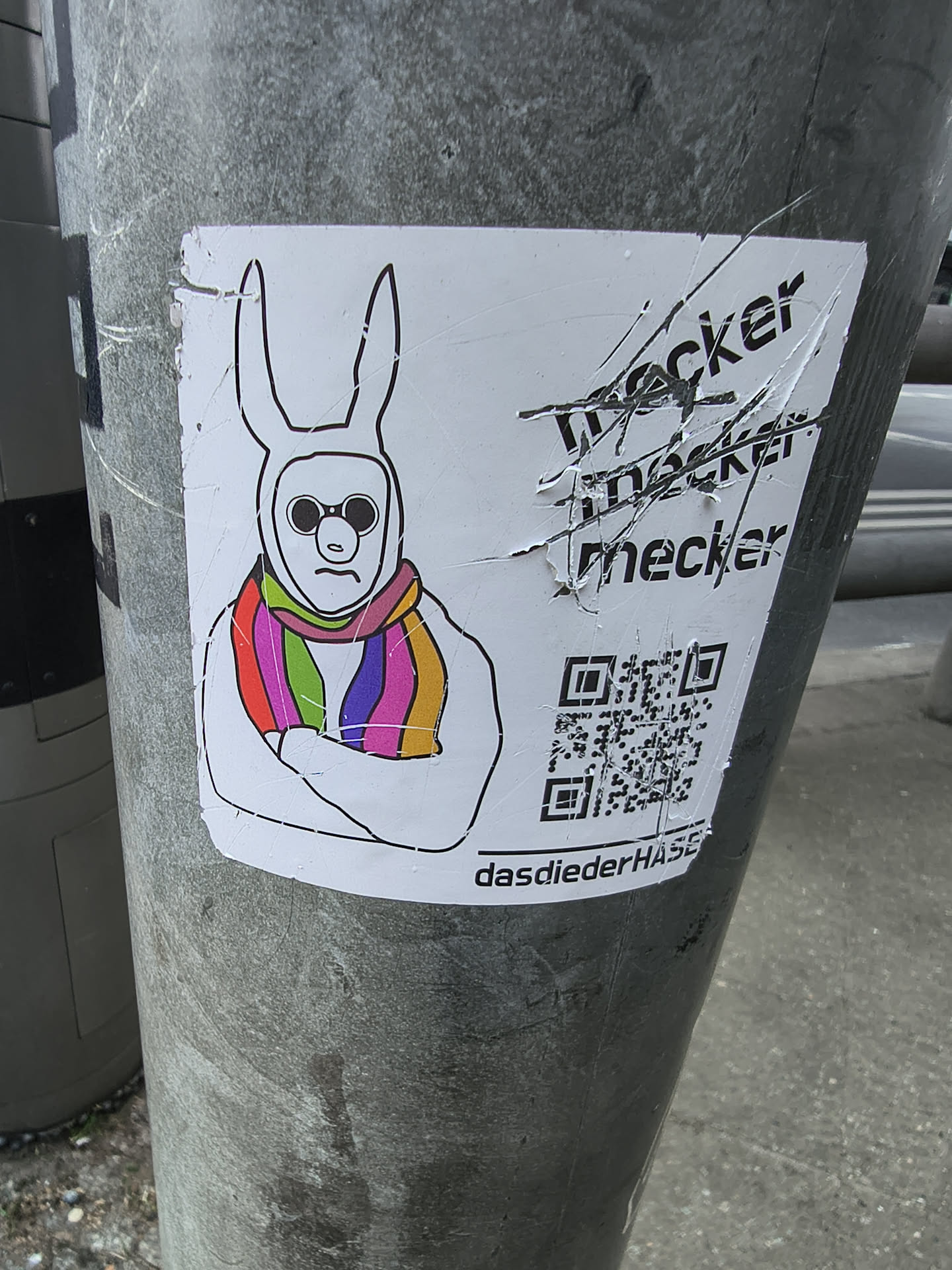 A white sticker featuring a stylized, somewhat minimalist drawing of a figure wearing a white bunny costume or mask with long ears. The face inside the mask has a simple, grumpy expression with a downturned mouth and round, dark sunglasses. The figure's arms are crossed over its chest. A prominent feature is a brightly colored rainbow-striped scarf wrapped around its neck, displaying vibrant bands of red, orange, yellow, green, blue, and purple. To the right of the bunny, there is black text that appears to be scratched out or crossed through, reading 'meckern' multiple times. Below this text is a QR code, and at the very bottom of the sticker, the text 'dasdiederHASE' is visible. The sticker itself shows signs of wear, including creases and slight peeling, indicating it's a physical sticker applied to a textured grey pole.