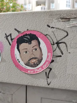 Street sticker A round pink sticker with an illustration of a man's face. The text on the sticker says "erst Pffi, dann Steffi". There are some small icons below the illustration, including Instagram and maybe other social media accounts.