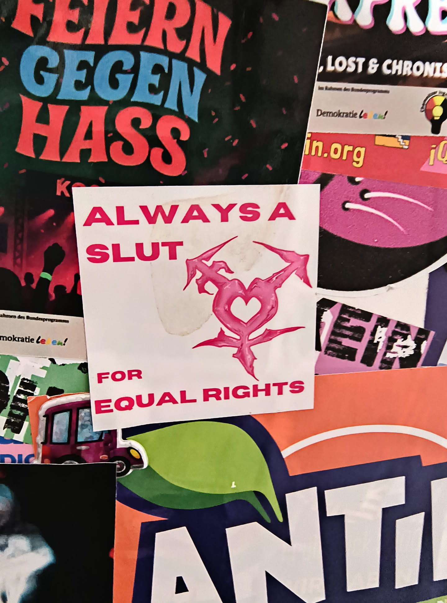 The sticker is square-shaped with a white background. It features pink text that reads 'ALWAYS A SLUT' at the top, and 'FOR EQUAL RIGHTS' at the bottom. In the center, there is a prominent pink graphic that combines the Venus symbol (female) with the Mars symbol (male) and an arrow, a common symbol representing transgender pride. The sticker appears to be slightly aged or has some smudges on its surface.