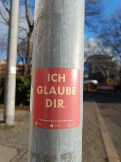 Street sticker A red sticker affixed to a metal pole.  The sticker features the German phrase "ICH GLAUBE DIR." (I believe you) in large, sans-serif lettering. Below this main text is smaller text indicating the sticker is part of a campaign against sexual abuse, along with what appear to be social media logos.