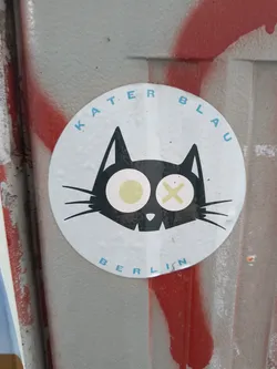 Street sticker The sticker is circular with a faded white background. Its central feature is a stylized black cat head with pointed ears and long whiskers. The cat's eyes are prominent, with the left eye (viewer's left) displaying a pale yellow circular pupil, while the right eye features a pale yellow 'X' shape. The mouth is depicted with a jagged, tooth-like line. Curving along the top edge of the circle, the words "KATER BLAU" are written in light blue capital letters. Similarly, along the bottom curve, "BERLIN" is written in the same light blue, uppercase font. The sticker appears slightly worn with minor edge peeling, set against a grey, textured background with visible red spray paint marks.