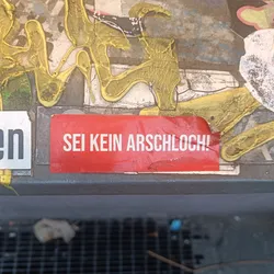 A red rectangular sticker with white text on it, attached to a surface with other graffiti and stickers. The text on the sticker is in German.