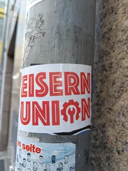 Street sticker A sticker featuring the text "EISERNE UNION" in bold red letters, accompanied by a red logo that seems to depict a gear with a wrench inside. The sticker is affixed to a gray metal pole.
