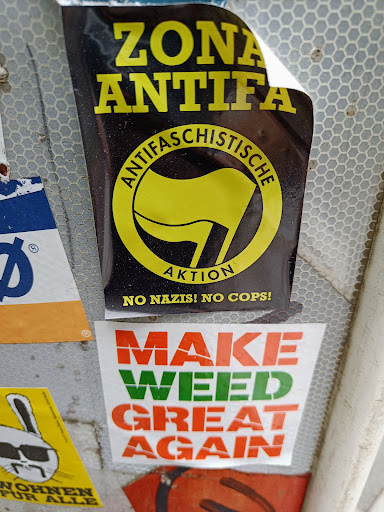 Street sticker A black sticker with yellow text and logo. The text includes "Zona Antifa",  "Antifaschistische Aktion", and "No Nazis! No Cops!". The logo is a yellow flag inside a circle.