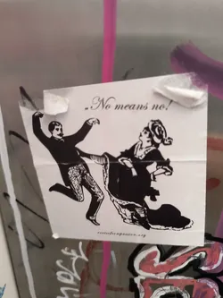 Street sticker The sticker is printed on white paper with black ink. It features a vintage-style illustration depicting a man in a suit kicking or gesturing forcefully towards a woman who is dressed in historical attire and appears to be recoiling or resisting. The text '„No means no!' is prominently displayed in a stylized font above the figures. Below the illustration, the URL 'rootsofcompassion.org' is printed in smaller text. The sticker is attached with visible pieces of tape and is slightly creased.