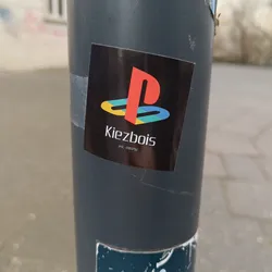 Street sticker Black square sticker with a stylized Playstation logo and the text 'Kiezbois' underneath.  It also has 'est. 2001/02' in smaller text below the name. The Playstation logo is in red, yellow, blue and green. The sticker appears to be on a gray pole.