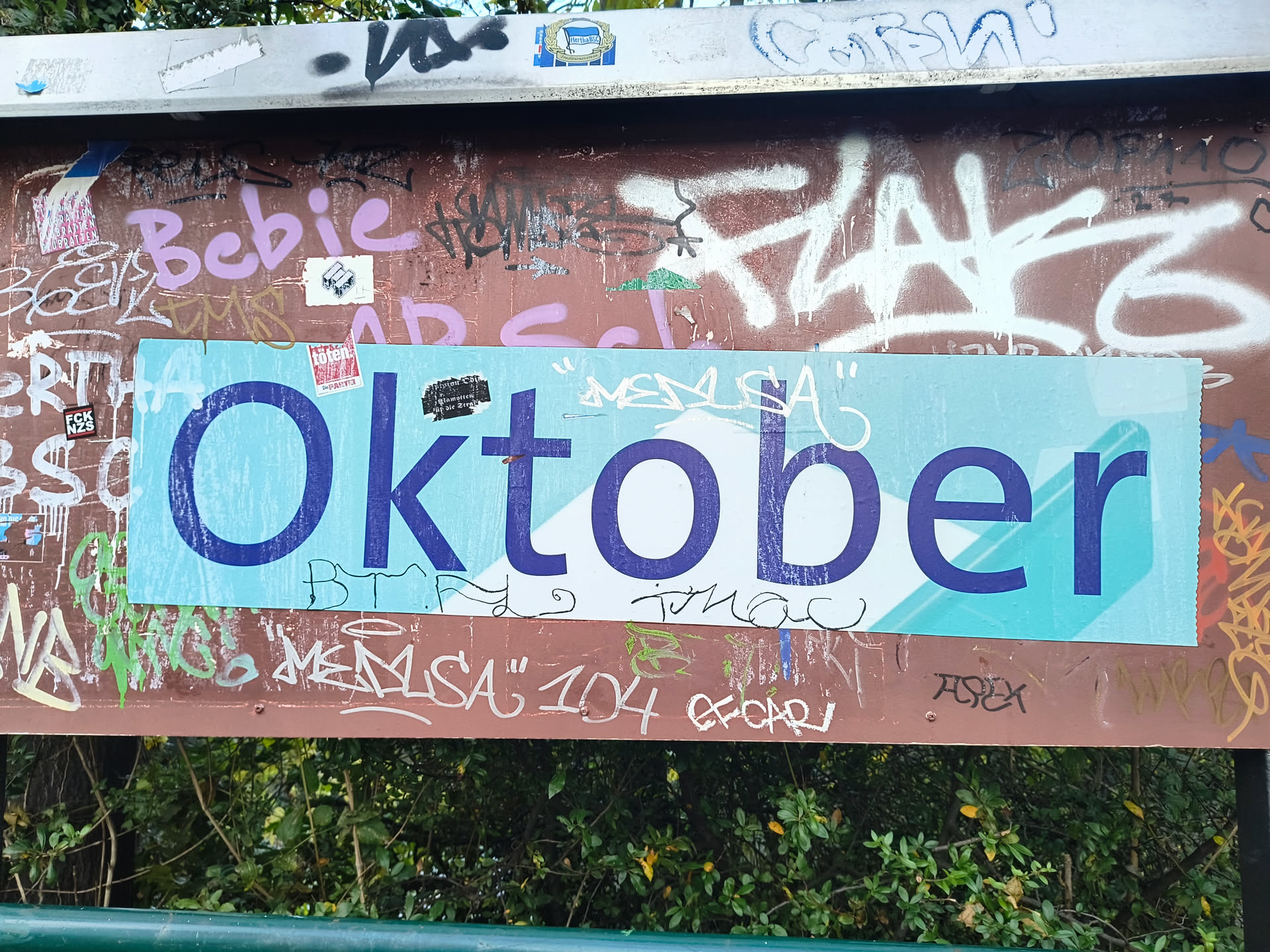 The central sticker is rectangular with a light blue background. It features the word 'Oktober' in large, bold, dark blue sans-serif letters. The letters are slightly distressed, with some areas appearing lighter or having subtle texture. The sticker is placed over a brown surface that is heavily covered in various graffiti tags and scribbles in different colors, including pink, white, black, and green. Some smaller stickers and pieces of paper are also visible around the main sticker.