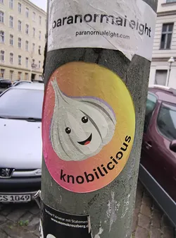 Street sticker The sticker is circular and features a stylized, cartoonish representation of a garlic bulb. The garlic has a light greyish-white color with subtle purple accents around the top cloves. It has two large, black, circular eyes and a wide, smiling mouth with a red tongue. The background of the sticker is a gradient that transitions from a bright yellow at the top to a pinkish-red at the bottom, with some subtle water droplets visible on the surface. The word 'knobilicious' is written in bold, black, sans-serif font along the bottom curve of the sticker.