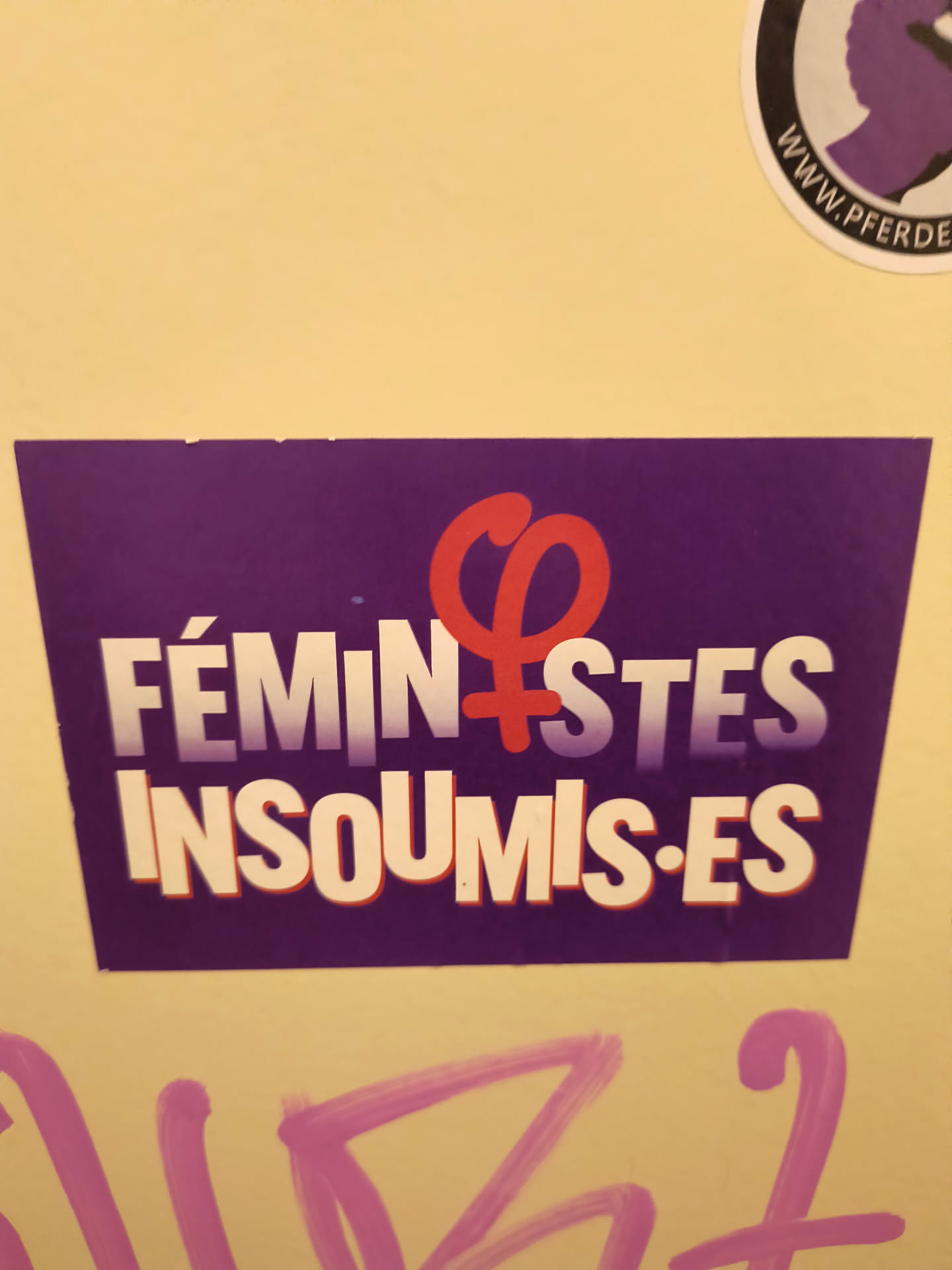 Street sticker The sticker is rectangular with slightly angled top corners, featuring a deep purple background. The main text, 'FÉMINISTES INSOUMIS·ES', is rendered in bold, white, sans-serif letters with a subtle red outline or shadow. A prominent, vibrant red stylized female gender symbol (Venus symbol) that also incorporates a raised fist replaces the 'I' in 'FÉMINISTES', symbolizing strength and resistance. The text 'INSOUMIS·ES' is positioned directly below 'FÉMINISTES'. The sticker is adhered to a light yellow wall, with some pink graffiti visible below it.