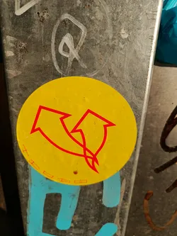 A round yellow sticker with two red upward arrows. The arrows are intertwined. The sticker is attached to a dirty gray metal surface.