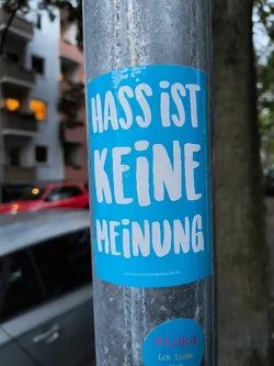 Street sticker The central sticker is rectangular with rounded corners and a bright blue background. It features large, white, sans-serif lettering that reads 'HASS IST KEINE MEINUNG' (Hate is not an opinion). Below this main text, in smaller white font, is the website 'www.zentrum-fur-demokratie.de'. To the right of the main text, in a vertical orientation and very small font, is additional text that appears to be contact or legal information. There is also a smaller, oval-shaped sticker at the bottom right of the main sticker, with pink and yellow text on a blue background, which reads 'minka, ich liebe dich'.