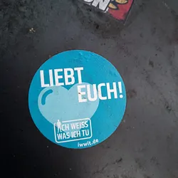Street sticker A round blue sticker with a stylized heart and the text "Liebt Euch!" (Love you!).  Smaller text below says "Ich weiss was ich tu" (I know what I'm doing) and a website address. 