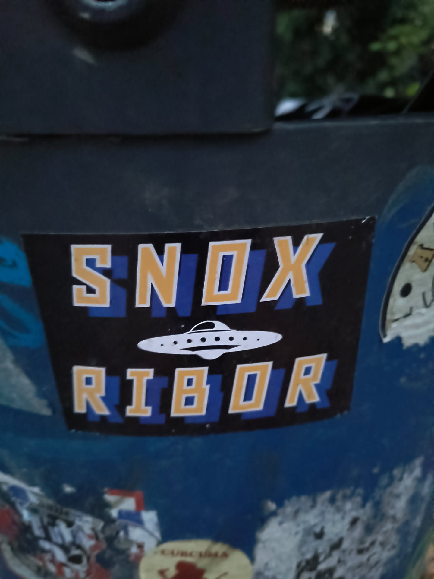 The sticker is rectangular with a black background. At the top, the word 'SNOX' is written in large, bold, yellow letters with a white outline, set against a blue background. Below 'SNOX', a white, stylized flying saucer with several windows is depicted. Beneath the UFO, the word 'RIBOR' is written in similar bold, yellow letters with a white outline, also against a blue background. The sticker appears to be slightly worn at the edges.