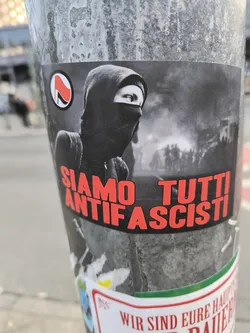 Street sticker A grayscale sticker featuring an image of a hooded person wearing a balaclava, possibly participating in a protest or demonstration, with text in red reading "Siamo Tutti Antifascisti."  A smaller symbol is visible in the upper left corner. The background features an out-of-focus image of a demonstration.