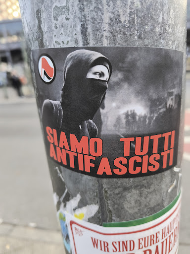 A grayscale sticker featuring an image of a hooded person wearing a balaclava, possibly participating in a protest or demonstration, with text in red reading "Siamo Tutti Antifascisti."  A smaller symbol is visible in the upper left corner. The background features an out-of-focus image of a demonstration.