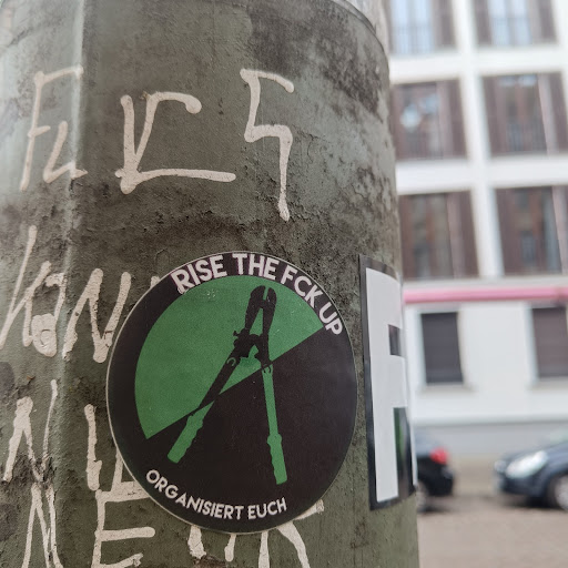 Street sticker A round sticker with a green and black half-circle design. It features a pair of pliers in green on a black background. The text "RISE THE FCK UP" is written in a semi-circle above the pliers, while "ORGANISIERT EUCH" is below. The sticker is affixed to a weathered gray pole, alongside other graffiti.