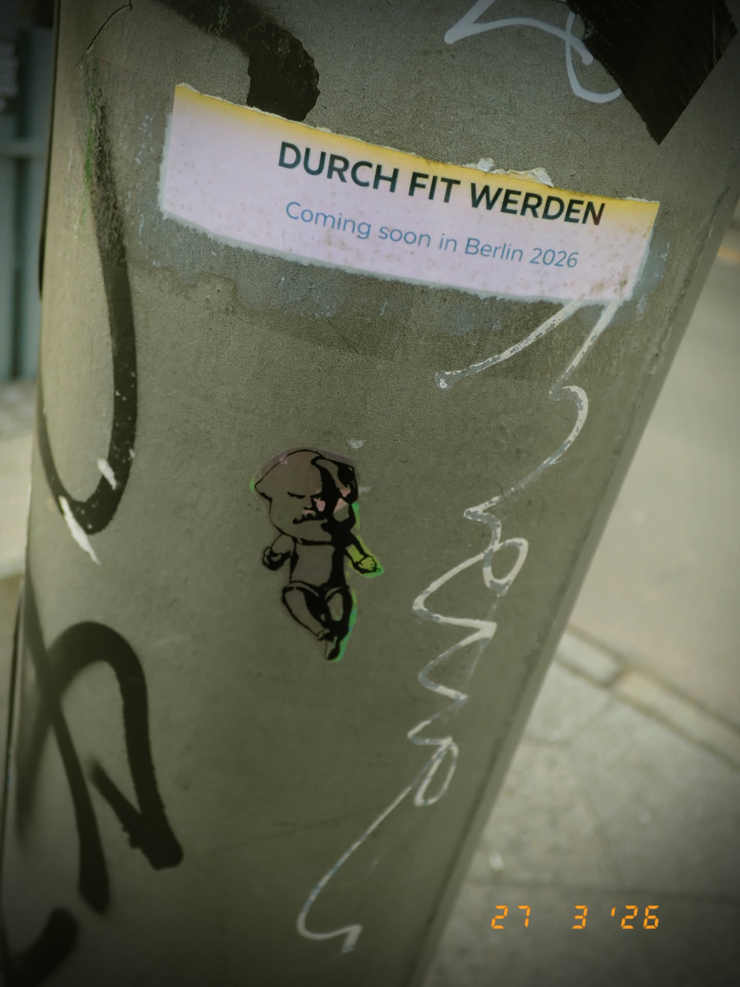 The sticker depicts a stylized illustration of a small, baby-like figure. The figure is predominantly dark grey or black, outlined in white, and features a vibrant, almost neon green and pink glow or aura around its edges, particularly on its left side. It has a determined or slightly mischievous expression, with small, dark eyes and a simple mouth. Its body posture suggests movement or readiness, with arms slightly bent and legs positioned as if walking or running. The overall style is cartoonish and graphic, affixed to a textured grey pole.