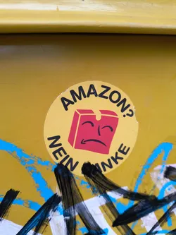Street sticker The sticker is circular with a yellow background. In the center, there is a red, cube-shaped object with a sad, downturned mouth and closed eyes, resembling a face. The text 'AMAZON?' is written in black capital letters in an arc above the box, and 'NEIN NICHT' is written in black capital letters in an arc below the box. The sticker appears to be slightly worn and has some minor tears around the edges. It is placed on a yellow surface that has been partially covered with graffiti in blue, black, and white.