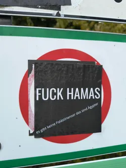 Street sticker The sticker is black with white text. The top line reads 'FUCK HAMAS' in large, bold, sans-serif capital letters. Below this, in smaller, italicized text, it says 'es gibt keine Palästinenser das sind Ägypter'. The sticker appears to be slightly torn on the left edge, revealing a white backing and a red circular shape behind it. The overall impression is one of a printed paper sticker applied to a surface.