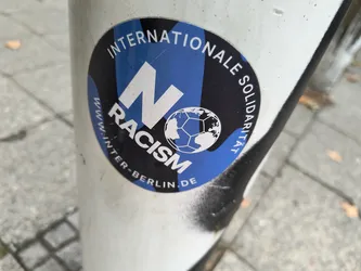 A round sticker with a blue and black design. It features a large 'N' over the word 'RACISM' in white, a stylized globe resembling a soccer ball, and the text 'Internationale Solidarität' in a circular arrangement.  The website www.inter-berlin.de is also visible.