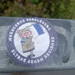 Street sticker A round sticker with a cartoon figure holding a flag. The text around the figure says "Nordkurve Babelsberg Ultras gegen Sexismus" which translates to "Nordkurve Babelsberg Ultras against Sexism". The sticker appears to be affixed to a railing outdoors.