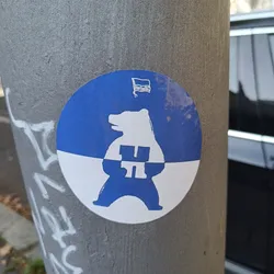 This round sticker is predominantly blue with a white bottom half. It depicts a bear standing on its hind legs, seemingly wearing or holding a house-shaped symbol on its chest. The bear and the house are depicted in white on the blue background. A small Ukrainian flag is visible in the top right corner of the sticker.