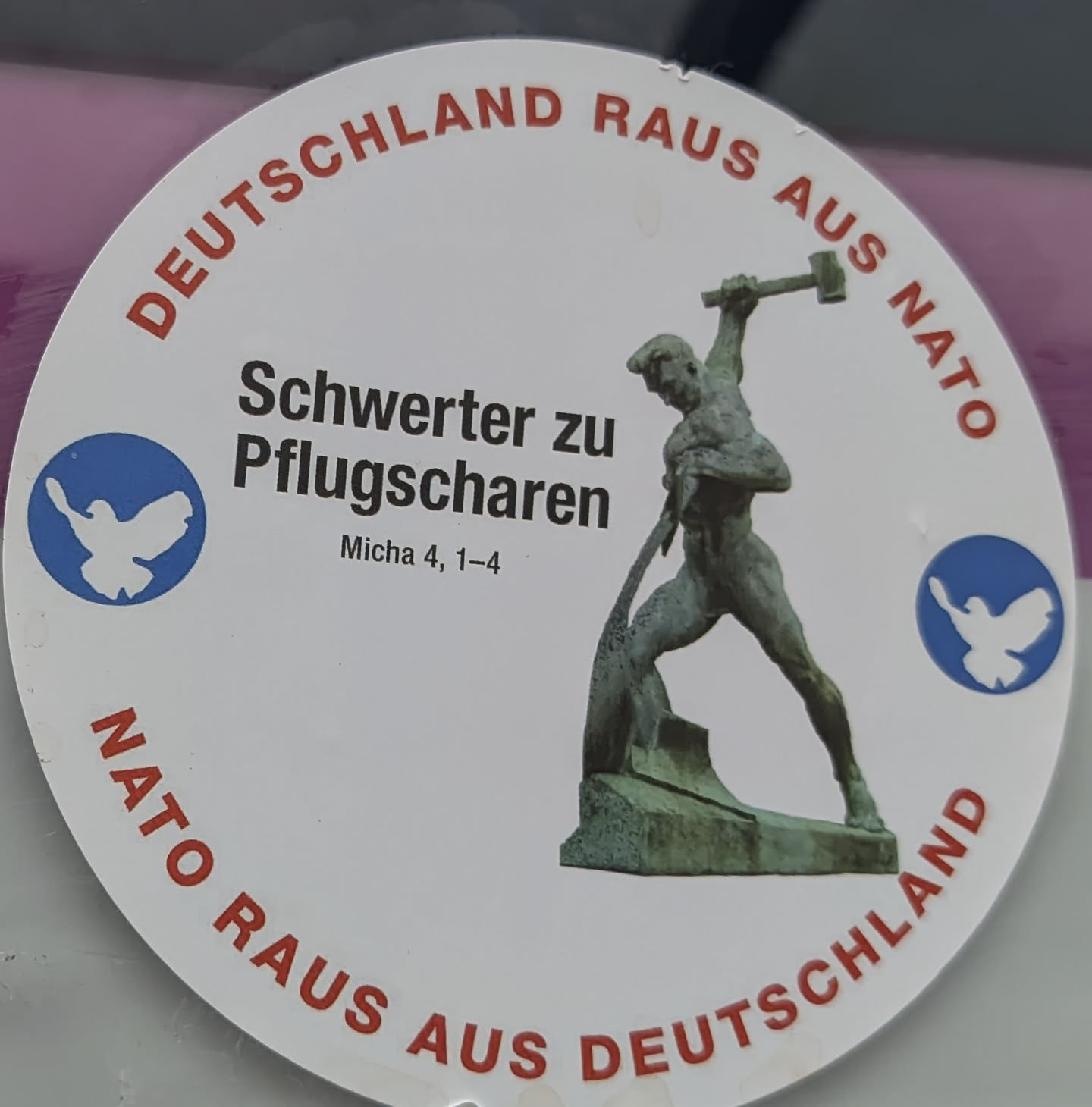 A circular white sticker featuring a central image of a bronze-colored statue depicting a muscular man forging a sword into a plowshare. To the left of the statue, in bold black text, are the words 'Schwerter zu Pflugscharen' (Swords into Plowshares), with a smaller biblical reference 'Micha 4, 1–4' below it. Flanking the central text and image are two blue circles, each containing a white silhouette of a dove, symbolizing peace. Curved around the top edge of the sticker in red capital letters is 'DEUTSCHLAND RAUS AUS NATO' (Germany out of NATO), and similarly curved around the bottom edge is 'NATO RAUS AUS DEUTSCHLAND' (NATO out of Germany). The overall design is clear, with strong contrasting colors and a distinct political and pacifist message.