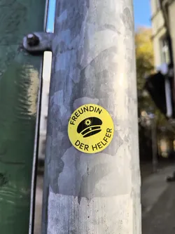 Street sticker A yellow circular sticker with black text and an illustration of a cap. The text says "Freundin der Helfer" which translates to "Friend of the Helpers" in German.