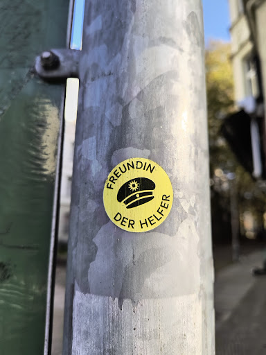 Street sticker A yellow circular sticker with black text and an illustration of a cap. The text says "Freundin der Helfer" which translates to "Friend of the Helpers" in German.