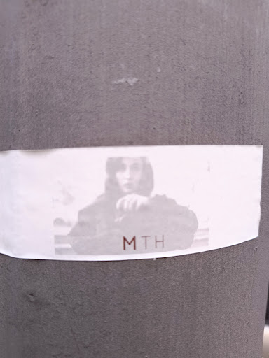 This sticker shows a person's photograph in grayscale with the text 'MTH' printed in reddish-brown font. The sticker is affixed to a gray, textured, cylindrical surface.