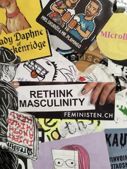 Street sticker The central sticker is a rectangular white sticker with black text. The top line reads 'RETHINK' in large, bold, sans-serif capital letters. Below it, 'MASCULINITY' is written in similar, but slightly smaller, bold capital letters. Beneath the 'MASCULINITY' text, the URL 'FEMINISTEN.CH' is printed in smaller, white capital letters on a black background bar that spans the width of the sticker. A partial view of a hand with manicured nails (teal and purple) is visible on the right side, touching the sticker.
