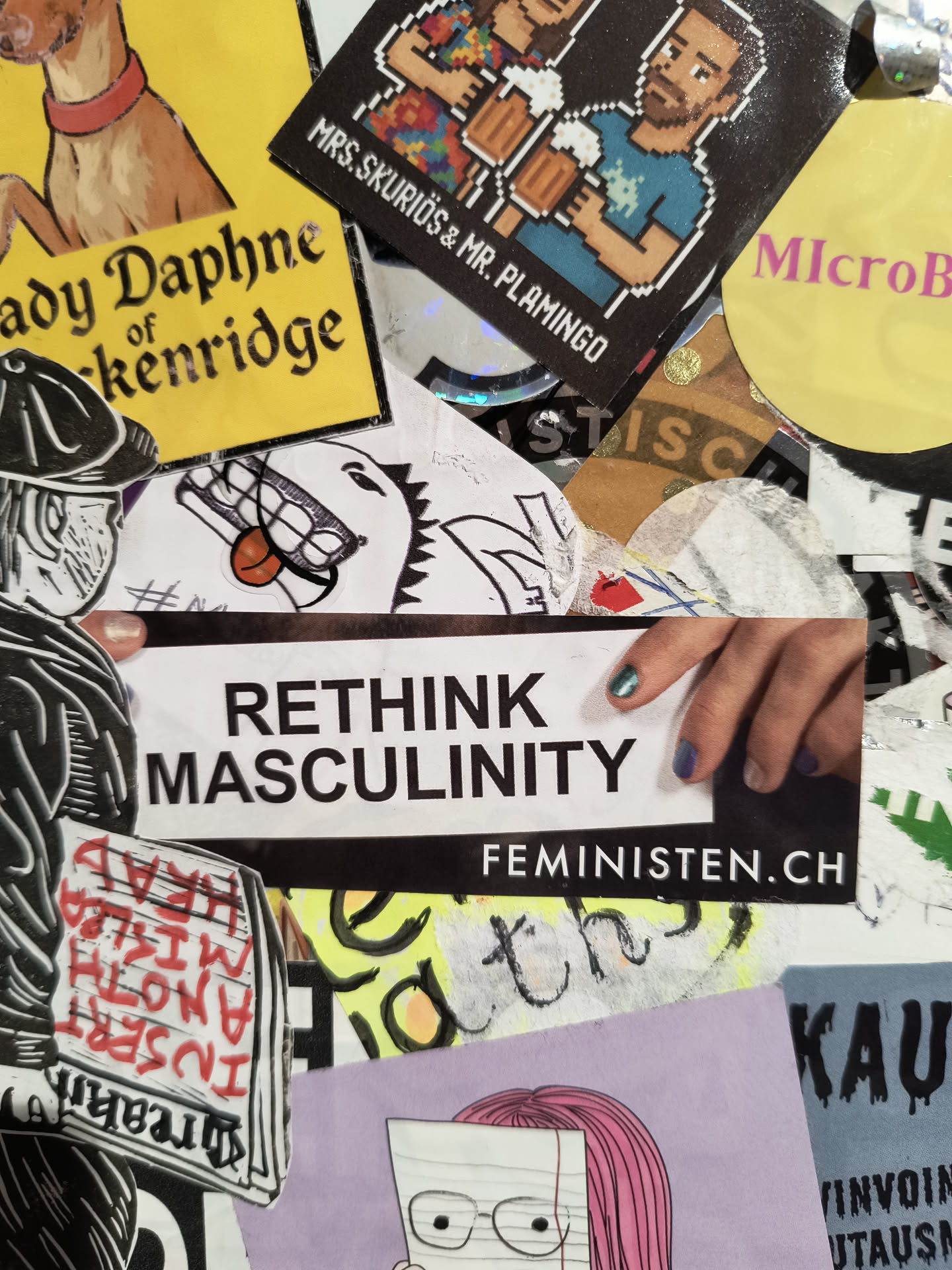 The central sticker is a rectangular white sticker with black text. The top line reads 'RETHINK' in large, bold, sans-serif capital letters. Below it, 'MASCULINITY' is written in similar, but slightly smaller, bold capital letters. Beneath the 'MASCULINITY' text, the URL 'FEMINISTEN.CH' is printed in smaller, white capital letters on a black background bar that spans the width of the sticker. A partial view of a hand with manicured nails (teal and purple) is visible on the right side, touching the sticker.
