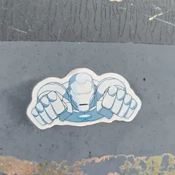 A sticker featuring Iron Man in a flying pose. The sticker is light blue and white, and Iron Man is depicted in a stylized cartoonish manner. The sticker is applied to a dark gray surface that looks like metal.
