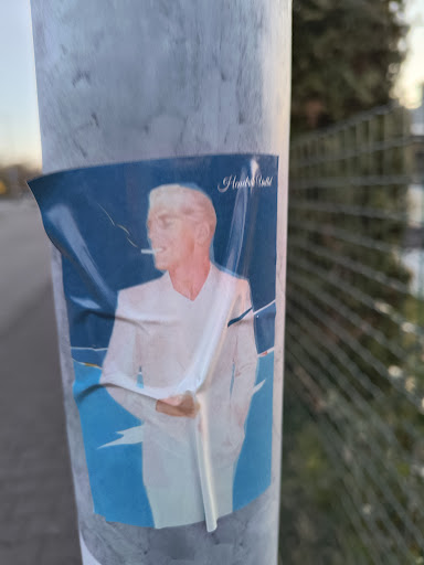 The sticker features a man with white hair and skin, smoking a cigarette. He is wearing a white suit against a blue background. The sticker is partially wrinkled and affixed to a metal pole.