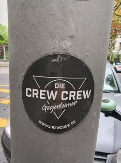 Street sticker The sticker is circular and predominantly black with white text and graphics. At the top, a white triangle points downwards. Below this, the words 'DIE CREW CREW' are prominently displayed in bold, sans-serif white capital letters. Beneath 'CREW CREW', the name 'Gegenbauer' is written in a flowing, cursive white font. At the bottom of the circle, the website 'WWW.CREWCREW.DE' is written in white, sans-serif capital letters. The sticker appears to be slightly worn or aged, with some minor scuffs and imperfections on its surface.